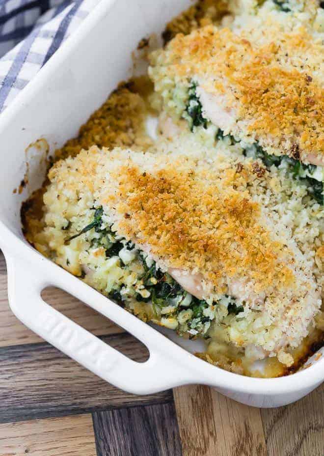 A white baking dish with handles, with stuffed and breaded chicken breasts inside it. Spinach stuffing is also visible inside the chicken breasts.