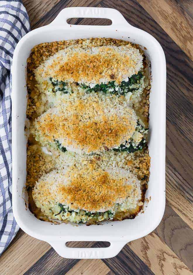 Overhead view of three breaded chicken breasts in a white rectangular baking dish. Chicken is stuffed feta and spinach.