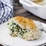 A breaded chicken breast on a double stacked white plate with a fork. Chicken is stuffed with feta and spinach. A baking dish and a blue and white linen are pictured in the background.