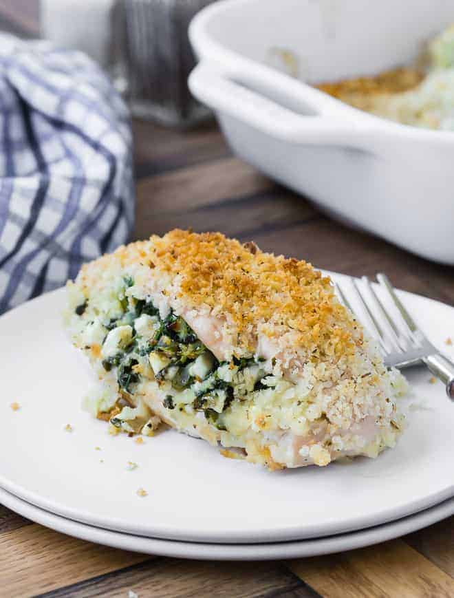 A breaded chicken breast on a double stacked white plate with a fork. Chicken is stuffed with feta and spinach. A baking dish and a blue and white linen are pictured in the background.