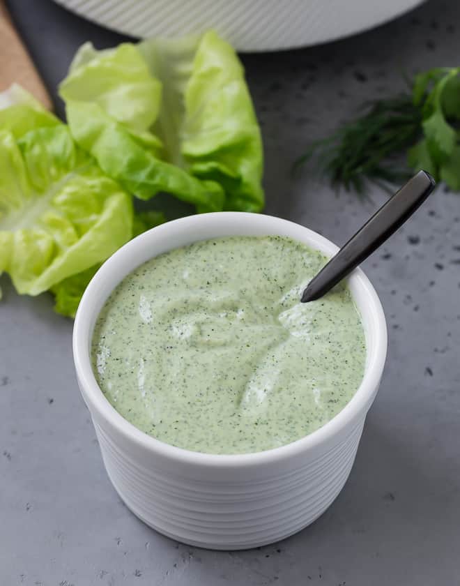 Overhead view of green goddess dressing in a small white bowl. A large bowl of salad is placed next to it. 