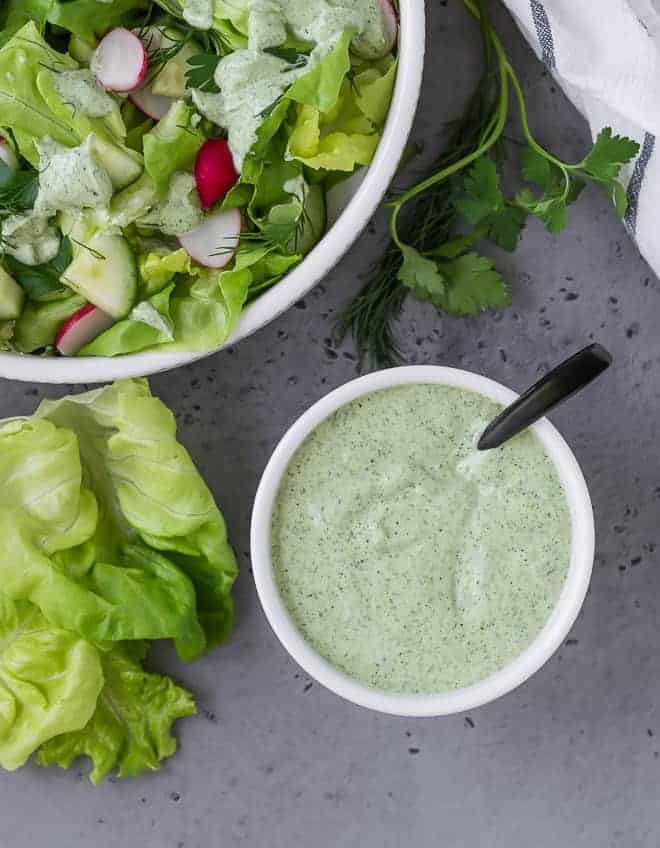 Overhead view of green goddess dressing in a small white bowl. A large bowl of salad is placed next to it. 