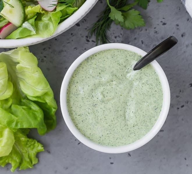 Overhead view of light green herby dressing in a small white bowl on a grey background.