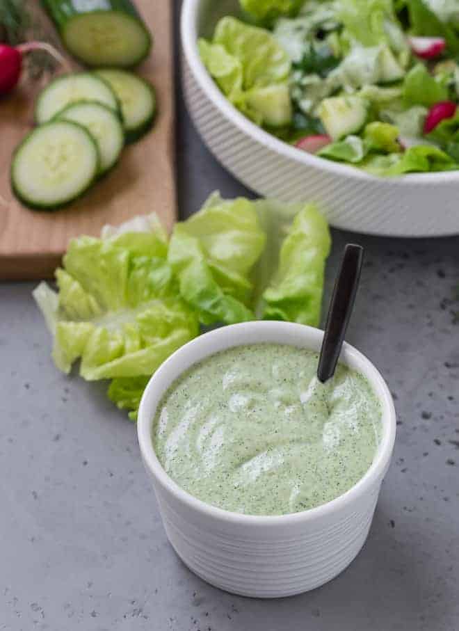 Green goddess dressing in a small white bowl. A large bowl of salad is placed next to it. 