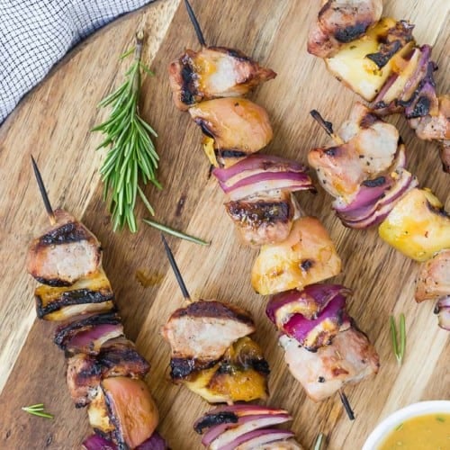 Overhead view of grilled pork, apple, and onion kabobs on a round wooden cutting board. A light orange sauce is also pictured in a small white bowl.