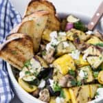 A white bowl filled with grilled zucchini, summer squash, mushrooms, and goat cheese. It is served with grilled bread pieces.