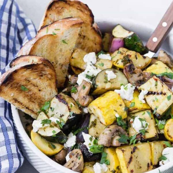 A white bowl filled with grilled zucchini, summer squash, mushrooms, and goat cheese. It is served with grilled bread pieces.