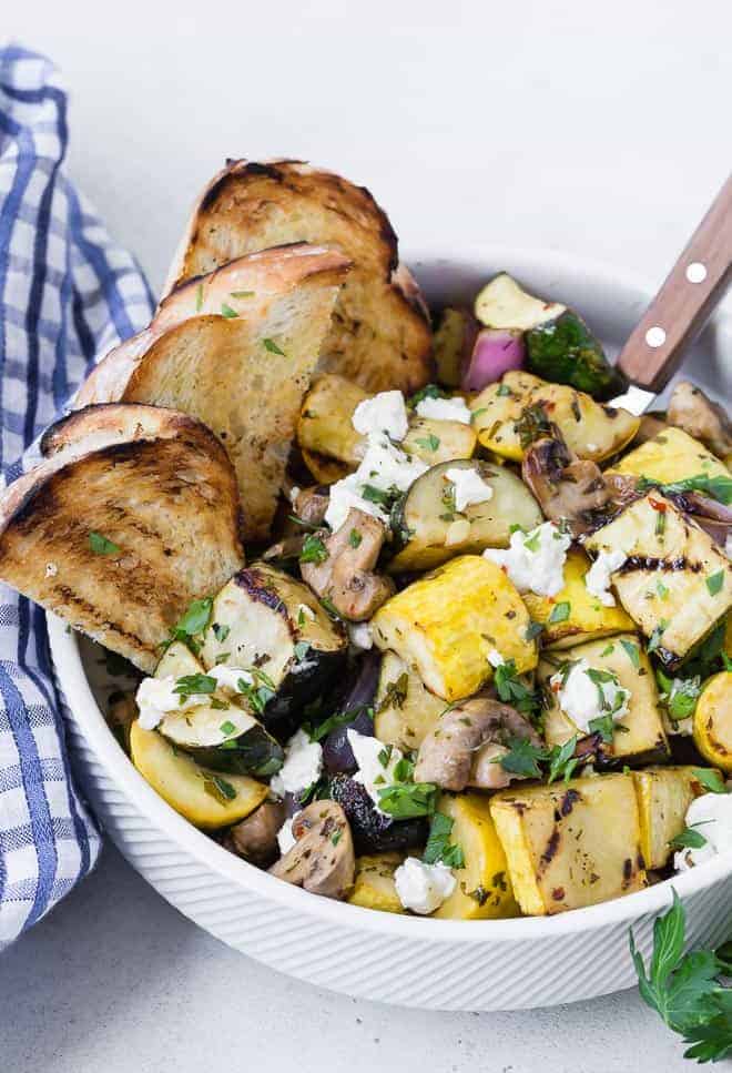 A white bowl filled with grilled zucchini, summer squash, mushrooms, and goat cheese. It is served with grilled bread pieces. 