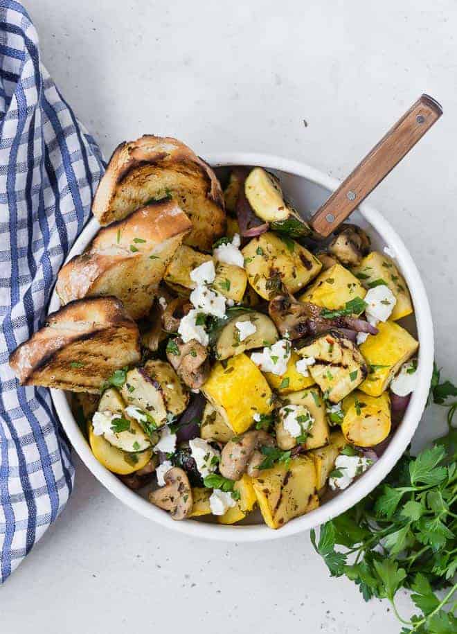 Overhead view of a white bowl on a light grey surface. Bowl is filled with grilled zucchini, summer squash, mushrooms, and goat cheese. It is served with grilled bread. A blue and white linen and fresh parsley is also pictured.