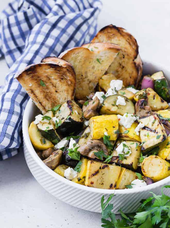 A white bowl on a light grey surface. Bowl is filled with grilled zucchini, summer squash, mushrooms, and goat cheese. It is served with grilled bread. A blue and white linen and fresh parsley is also pictured.