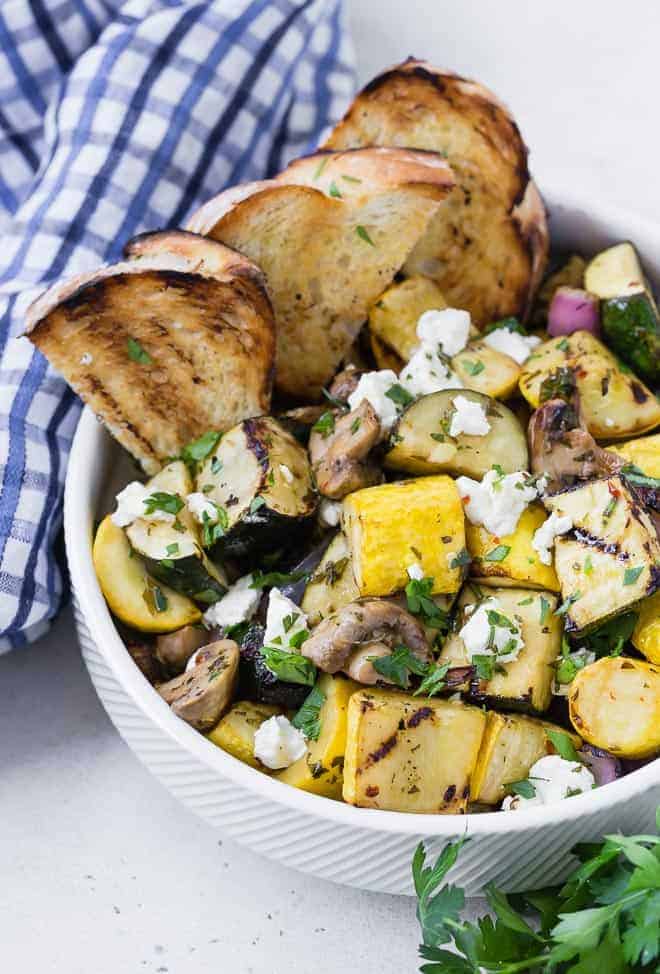 A white bowl on a light grey surface. Bowl is filled with grilled zucchini, summer squash, mushrooms, and goat cheese. It is served with grilled bread. A blue and white linen and fresh parsley is also pictured.