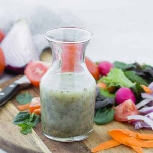 Small glass bottle of homemade italian dressing on wooden cutting board surrounded by colorful vegetables.