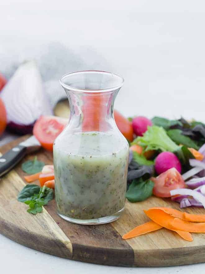 Small glass bottle of homemade italian dressing on wooden cutting board surrounded by colorful vegetables.