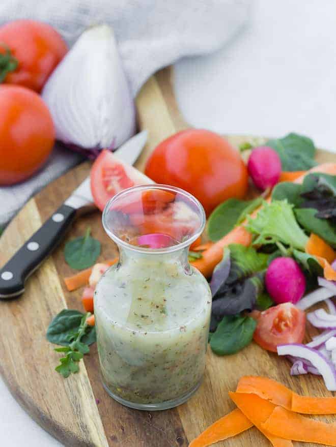 Small glass bottle of homemade italian dressing on wooden cutting board surrounded by colorful vegetables.