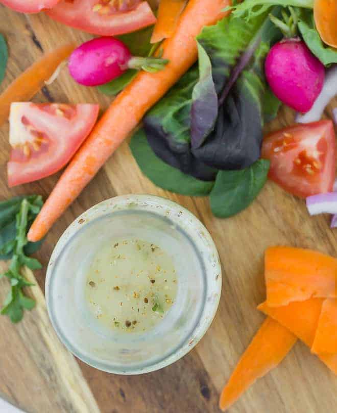 Overhead view of Italian dressing in a small glass bottle, surrounded by fresh vegetables on a wooden cutting board.