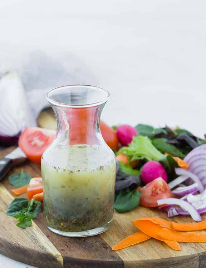 Small glass bottle of homemade italian dressing on wooden cutting board surrounded by colorful vegetables.