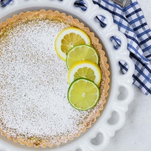 Overhead view of a tart on a white plate. The tart is topped with lots of powdered sugar, and lime and lemon wheels.