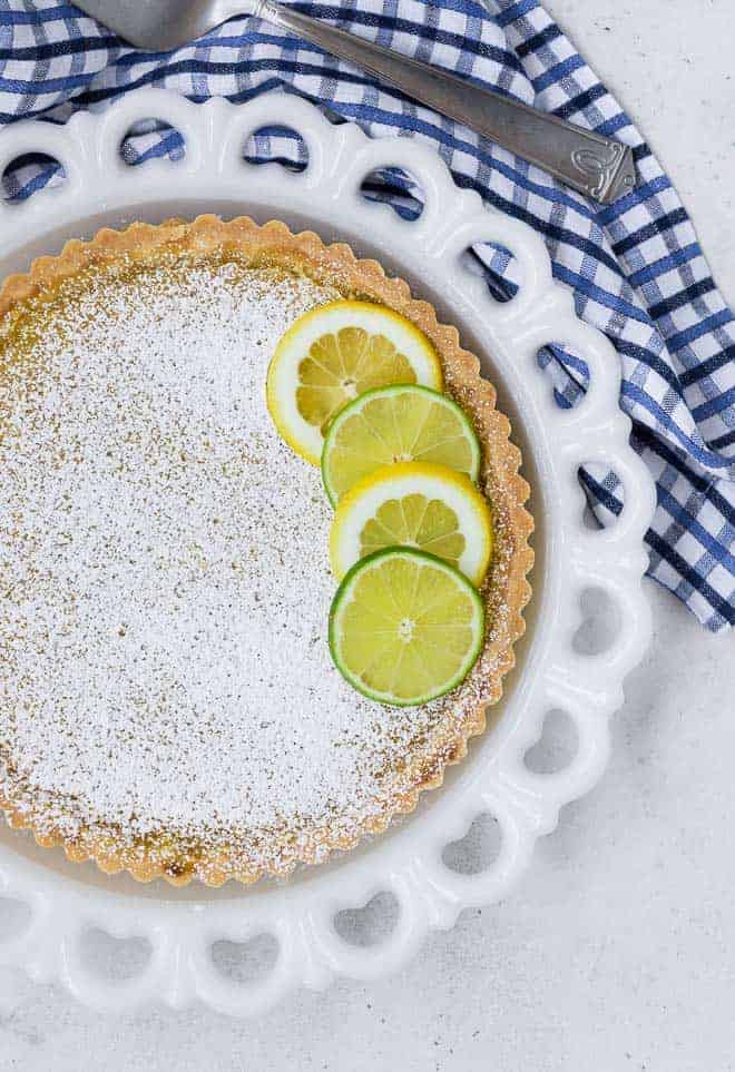 Overhead view of a tart on a white plate. The tart is topped with lots of powdered sugar, and lime and lemon wheels.
