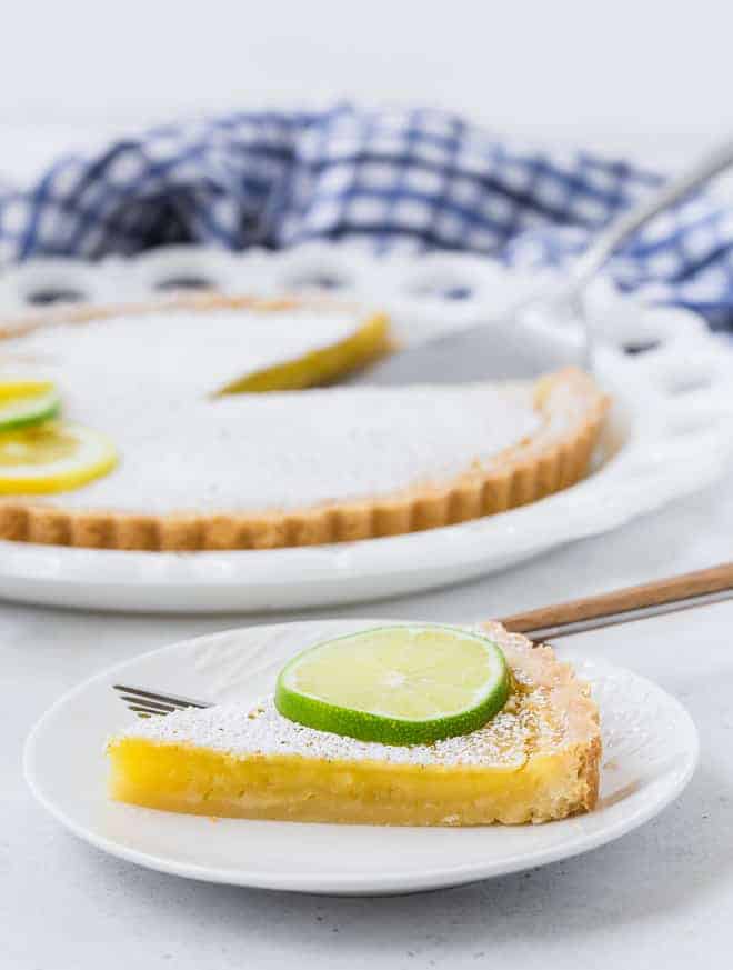 A slice of lemon lime tart on a plate, with the rest of the tart in the background.