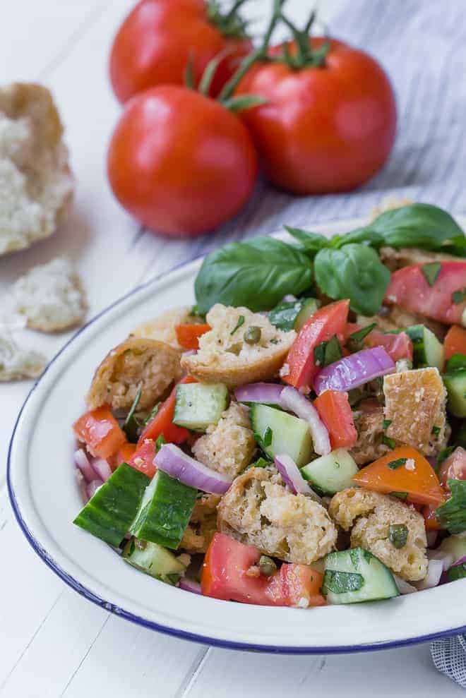 A summer salad on a large white and blue platter. Salad includes chopped tomatoes, cucumbers, fresh herbs, red onion, torn bread, and capers.