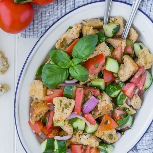 Overhead view of a panzanella salad on a large white and blue platter. Salad includes chopped tomatoes, cucumbers, fresh herbs, red onion, torn bread, and capers.