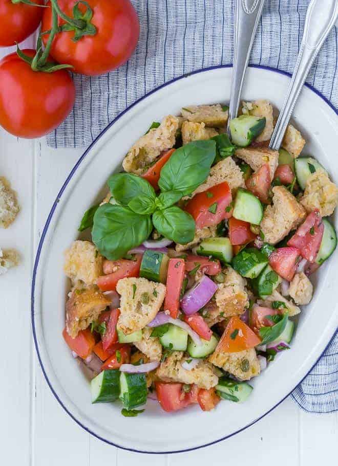 Overhead view of a panzanella salad on a large white and blue platter. Salad includes chopped tomatoes, cucumbers, fresh herbs, red onion, torn bread, and capers.