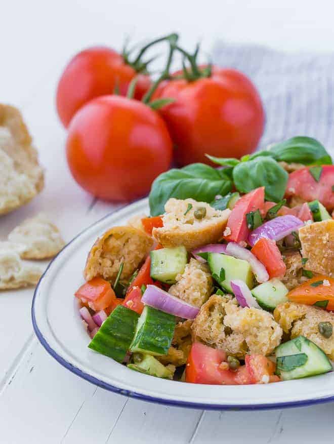 A summer salad on a large white and blue platter. Salad includes chopped tomatoes, cucumbers, fresh herbs, red onion, torn bread, and capers. Vine-ripened tomatoes and more torn bread are pictured in the background.