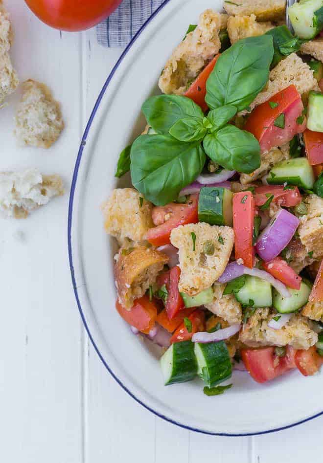 Overhead view of a panzanella salad on a large white and blue platter. Salad includes chopped tomatoes, cucumbers, fresh herbs, red onion, torn bread, and capers.