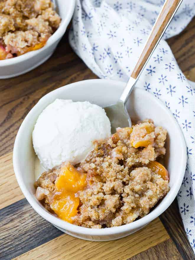 Overhead view of peach cobbler in a round white bowl, topped with ice cream. Another plate is partially visible. Both are on a wooden surface, and a blue and white linen is also pictured.