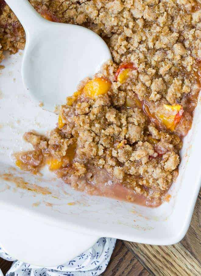 A white baking dish full of peaches and a crisp brown topping, being scooped out with a white ceramic spoon.