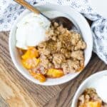 Overhead view of peach cobbler on a round white plate, topped with ice cream. Another plate is partially visible. Both are on a wooden surface, and a blue and white linen is also pictured.