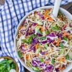 Overhead view of vinegar coleslaw in a bowl with a spoon in it. A small bowl of sliced green onions is also pictured, as well as a blue and white checkered cloth.