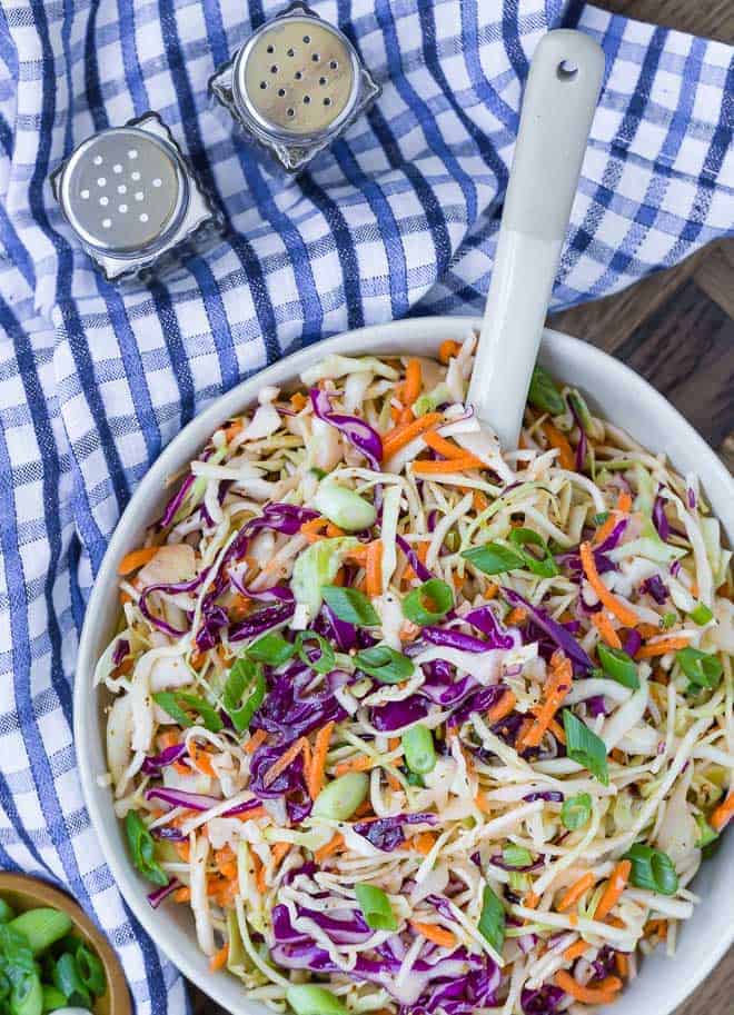 Overhead view of coleslaw in a bowl with a spoon in it. A small bowl of sliced green onions is also pictured, as well as a blue and white checkered cloth.