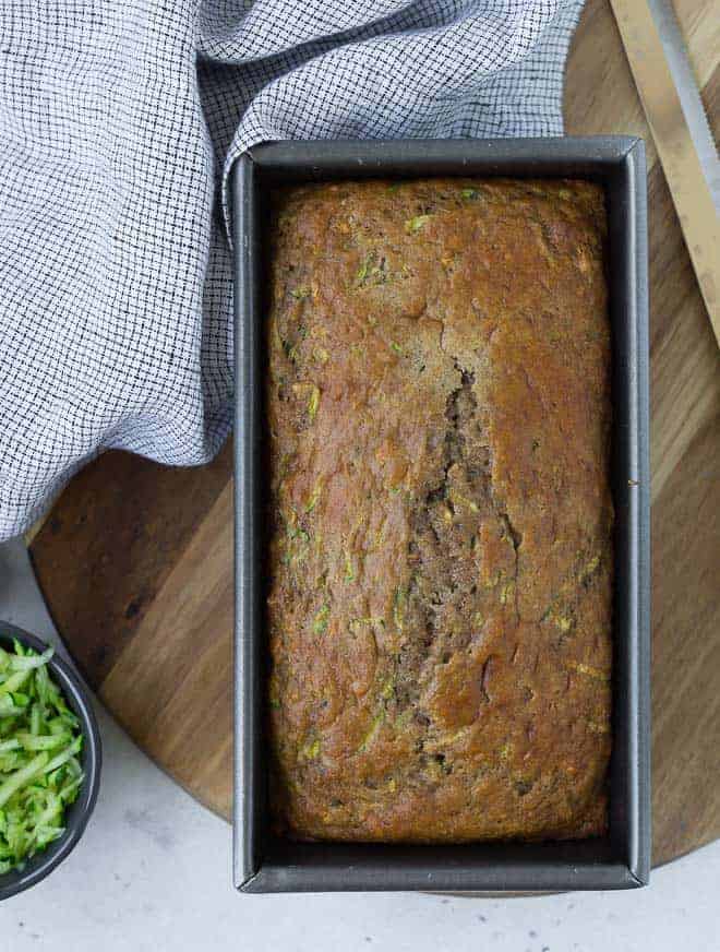 Overhead view of a loaf of bread in a metal loaf pan. A small bowl of shredded zucchini sits next to it.