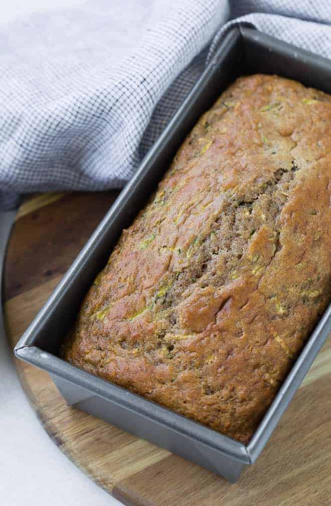 A silver loaf pan on a wooden surface, with a loaf of zucchini bread in the pan.