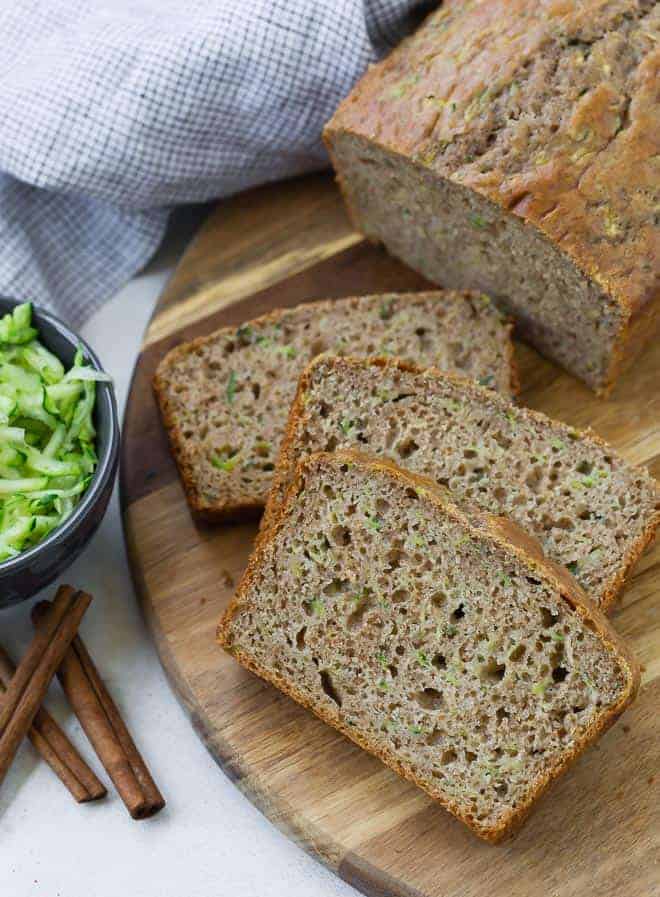 Sliced zucchini bread on a wooden surface. A small bowl of shredded zucchini sits next to the bread, as well as three cinnamon sticks.