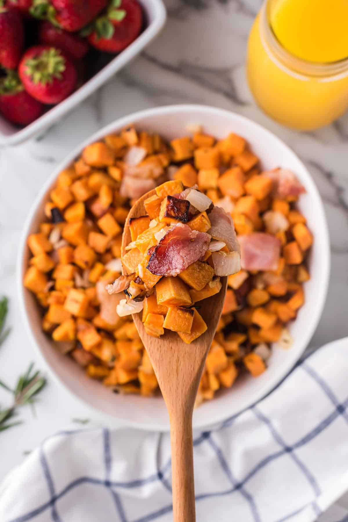 Air fried sweet potatoes, bacon, and onions on a wooden spoon over a white bowl full of mixture.