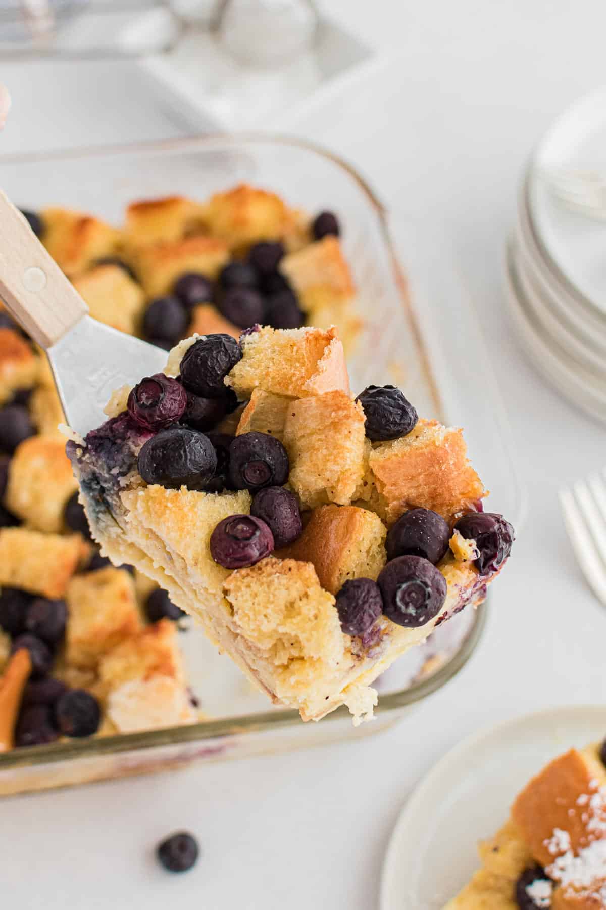 A slice of blueberry bread pudding being scooped out of a square glass pan.