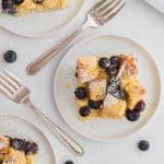 Overhead view of three slices of blueberry bread pudding on round white plates. Forks and fresh blueberries are also pictured.