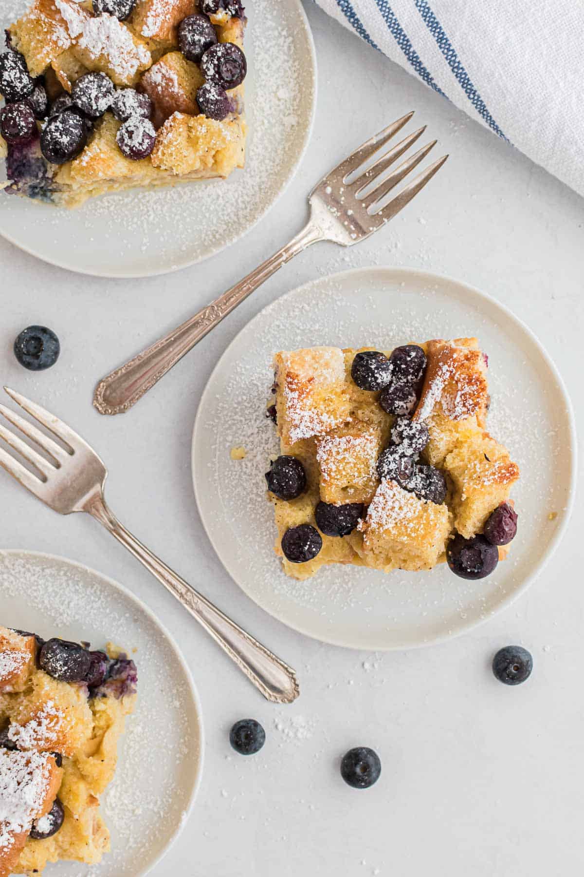 Overhead view of three slices of blueberry bread pudding on round white plates. Forks and fresh blueberries are also pictured.