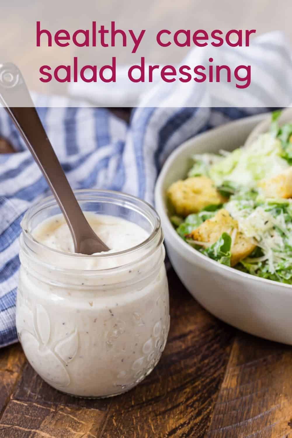 A jar of caesar salad dressing in front of a tossed salad. A text overlay reads "healthy caesar salad dressing."