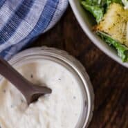 A jar of caesar salad dressing in front of a tossed salad. A text overlay reads "healthy caesar salad dressing."