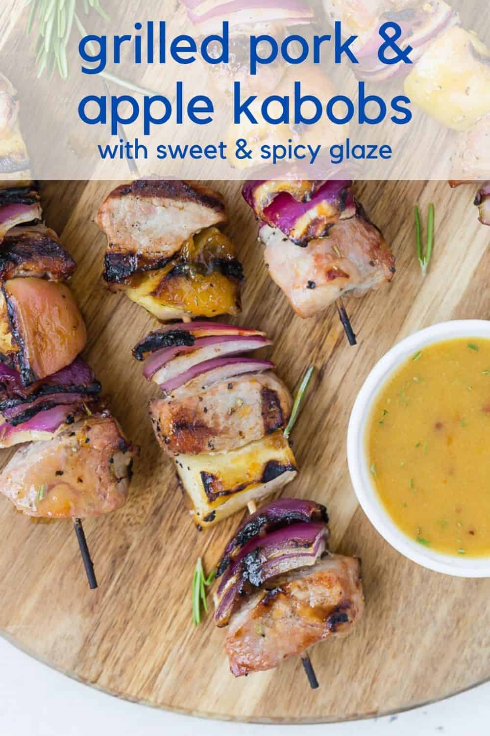 Kabobs of pork chunks, apples, and onions sit on a wooden cutting board. A light orange dipping sauce is in a white bowl next to them. Text overlay reads, "grilled pork & apple kabobs with sweet & spicy glaze."