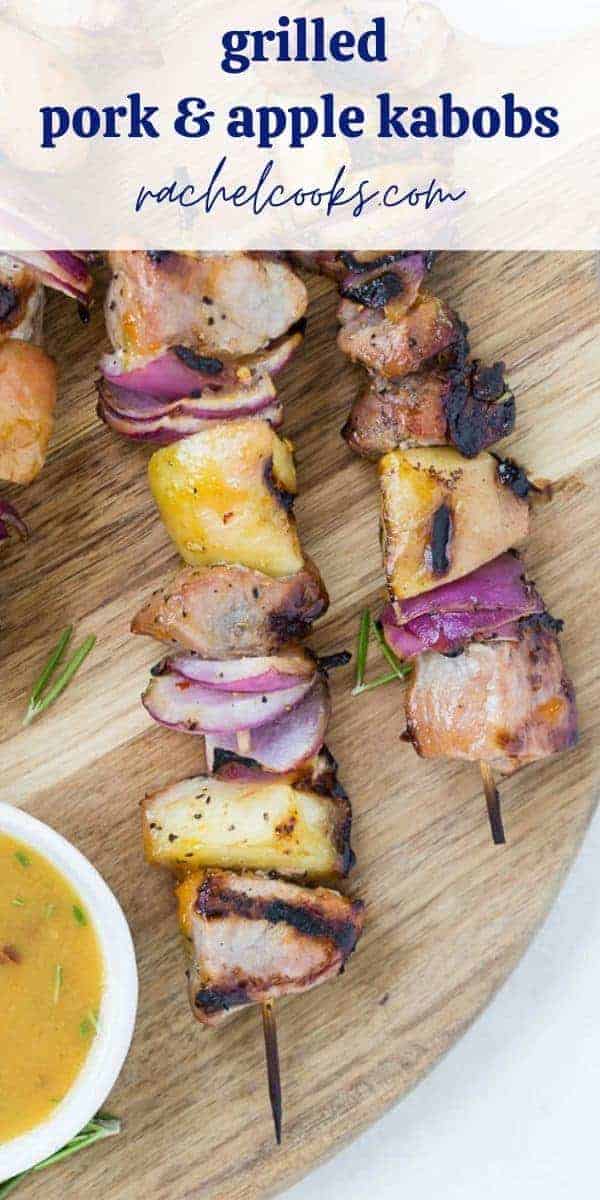 Kabobs of pork chunks, apples, and onions sit on a wooden cutting board. A light orange dipping sauce is in a white bowl next to them. Text overlay reads, "grilled pork & apple kabobs with sweet & spicy glaze."