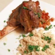 A white plate with a lamb shank, topped with a tomato based sauce and served with couscous, garnished with parsley.