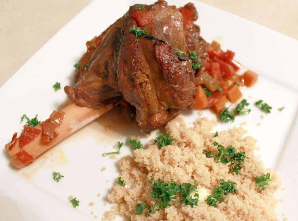 A white plate with a lamb shank, topped with a tomato based sauce and served with couscous, garnished with parsley.