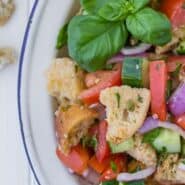 Salad on a white plate, consisting of tomatoes, cucumbers, croutons, fresh herbs, and red onions. Text overlay reads "How to Make Panzanella."