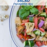 Salad on a white plate, consisting of tomatoes, cucumbers, croutons, fresh herbs, and red onions. Text overlay reads "the best panzanella salad - rachelcooks.com"