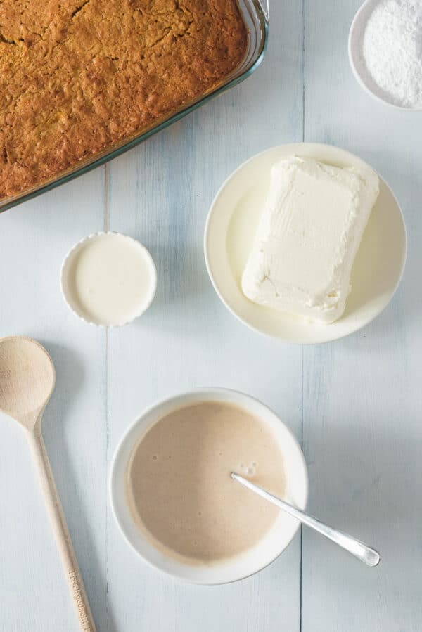 Cinnamon condensed milk in a bowl for the poke cake filling, next to a baked cake and more ingredients.