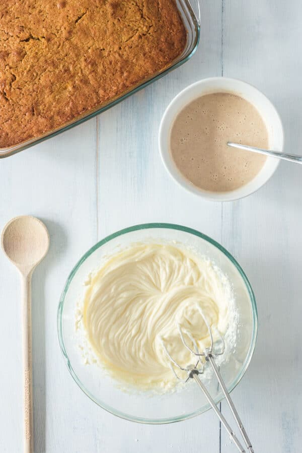 Cream cheese frosting ingredients combined in a bowl next to a smaller bowl of cinnamon condensed milk and a baked pumpkin cake.